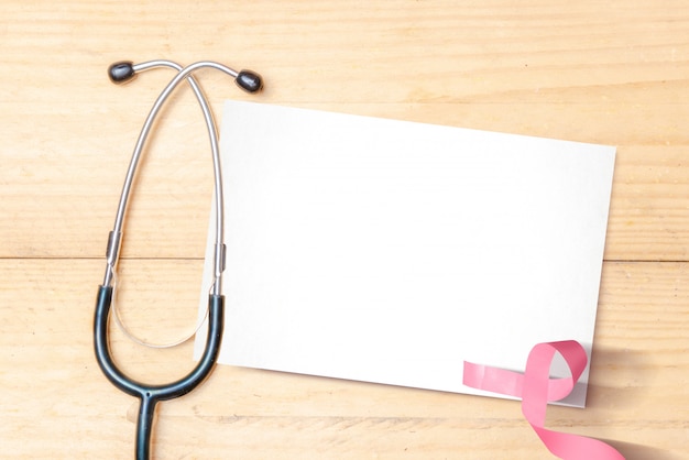 Pink awareness ribbon and stethoscope with empty white paper on a wooden