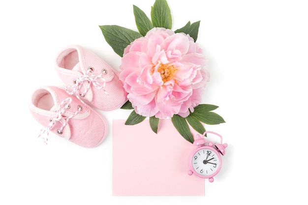 Pink baby girl shoes with peony flower alarm clock and a blank card for message on a white background Newborn greeting card or invitation Copy space Flat lay