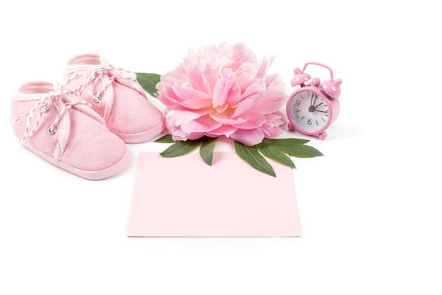 Pink baby girl shoes with peony flower alarm clock and a blank card for message on a white background Newborn greeting card or invitation Copy space