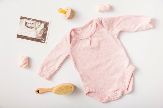 Photo pink baby onesie with sonography picture; pacifier; marshmallow; brush on white background