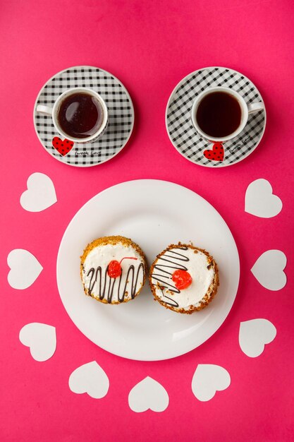 On a pink background cakes in a plate decorated with paper hearts and two cups of coffee top view