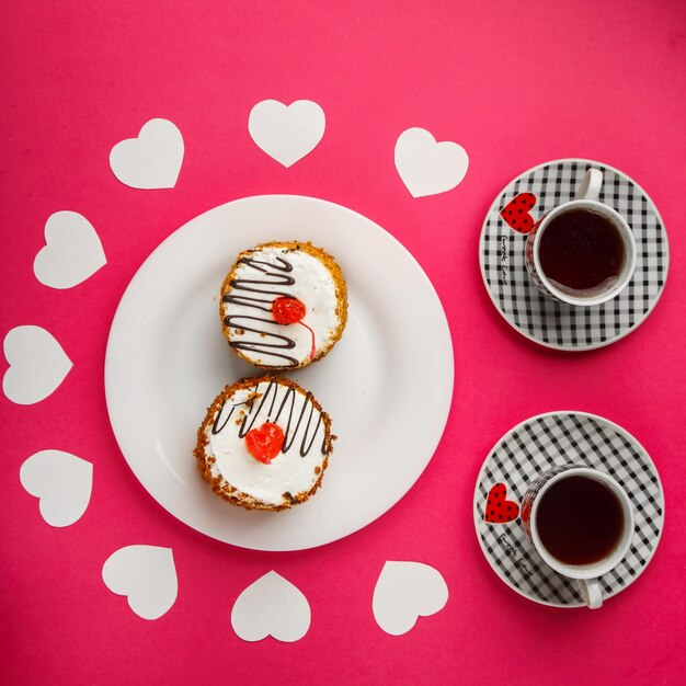 On a pink background cakes in a plate decorated with paper hearts and two cups of coffee