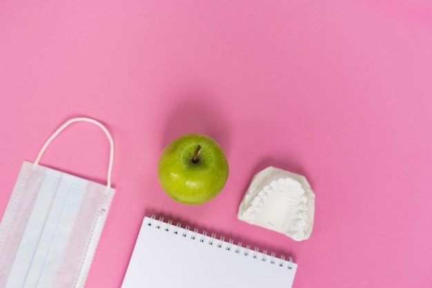 On a pink background is a cast of teeth, an apple, and a medical mask