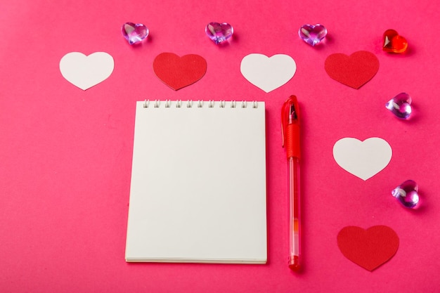 분홍색 배경에 빨간색과 흰색 하트와 발렌타인 데이용 펜이 있는 메모장