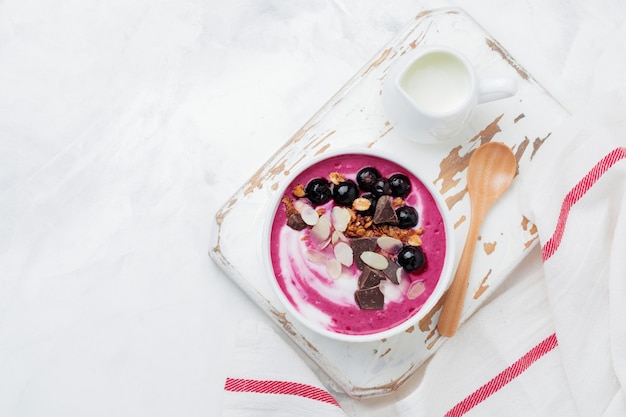 Pink black currant berries, banana smoothie bowl with granola, flakes from almonds and chocolate on light gray concrete surface