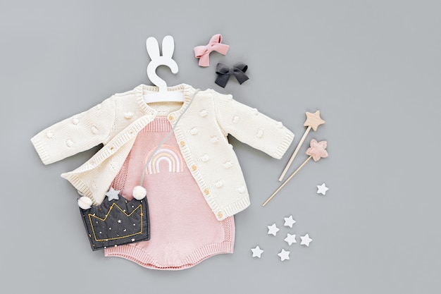 Photo pink bodysuit with knitted jumper, kids handbag shape of crown on cute hanger with bunny ears. set of  baby clothes and accessories  on gray background. fashion childs outfit. flat lay, top view