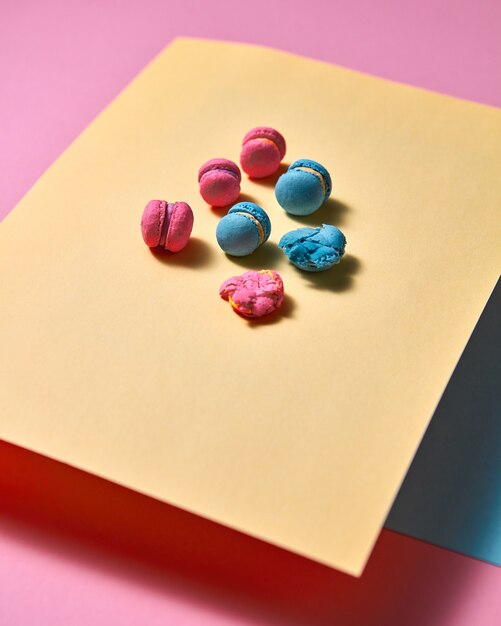 Pink bright macaroons are presented in the form of a triangular pyramid on yellow cardboard and one crushed macaroon on a pink paper background with reflection of shadows. Creative composition