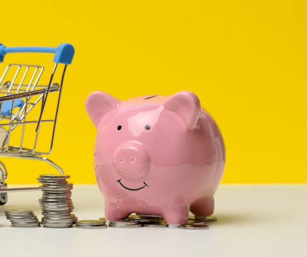 Pink ceramic piggy bank and a stack of coins on a white table, yellow background. Savings concept, budget planning. Income growth