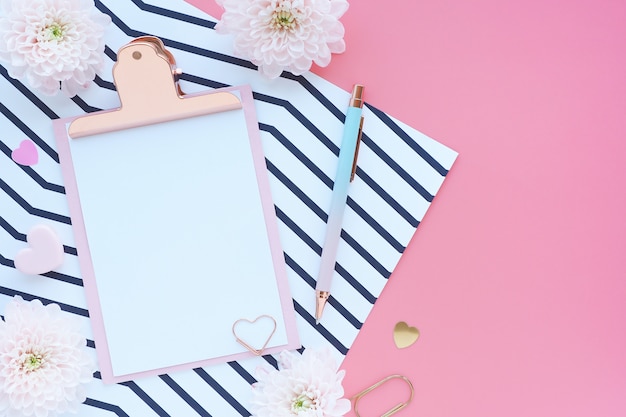 pink clipboard, pen, paper clip, striped paper and chrysanthemum flowers on a pink table.  Flat layout, top view.