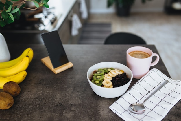 Pink coffee cup, bowl with chopped tropical fruits kiwi and banana, blueberries, spoon and mobile phone on bar counter in stylish loft kitchen.