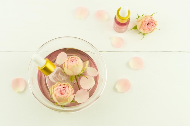 Розовая косметическая вода в миске с лепестками роз, бутылочки-капельницы с экстрактом роз на белом фоне. вид сверху.