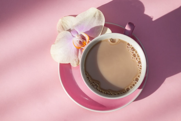 Pink cup of coffee and orchid flower on pink background. Top view.