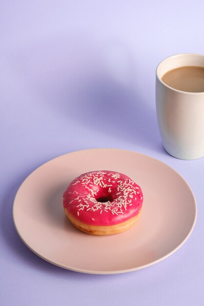 Pink donut with sprinkles on plate near to cup of coffee, sweet glazed dessert food and hot drink on blue minimal background, angle view