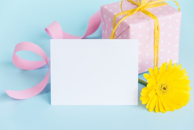 Pink dotted gift box, yellow gerbera flower and empty card over blue.