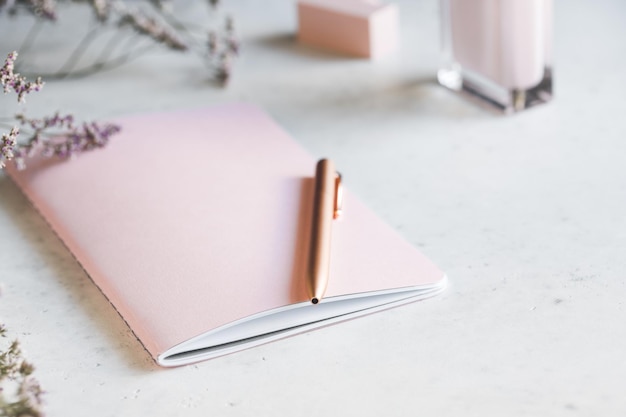 Pink empty paper notebook with golden pen on white table surrounded by flowers and fragrance The concept of femininity workplace