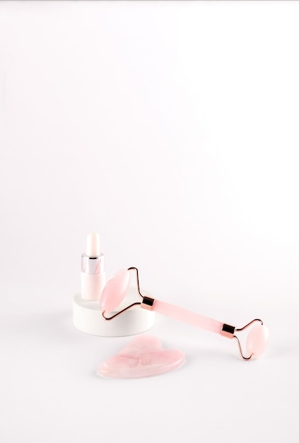 Pink face roller and gua sha massager made from natural quartz  stone with cosmetic massage oil over white background.  Lifting and toning treatment at home.