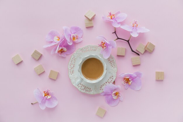 Pink gift bag and flying orchid flowers on light pink surface. Top view greeting card with delicate flowers, cup of coffee and empty wooden blocks. Women's day, mother's day greeting concept.