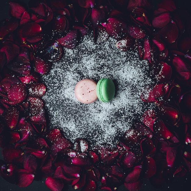 Pink and green macaroon lie on white powder in the circle of red rose petals