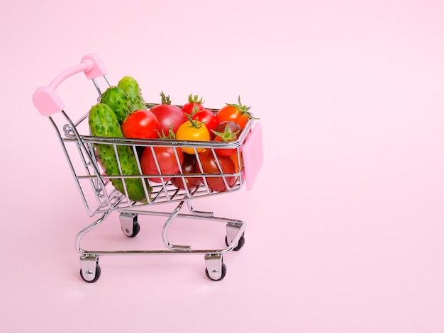 Розовая продуктовая тележка с помидорами и огурцами на розовом фоне