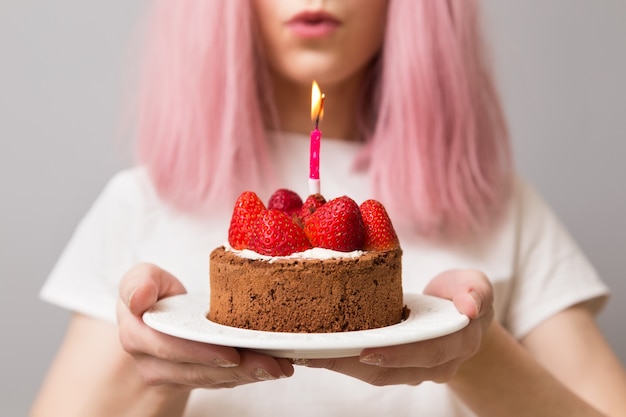 Девушка с розовыми волосами держит в руках свечу клубничный торт на день рождения.