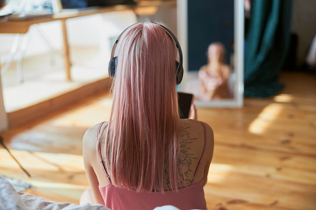 Photo pink haired woman with tattoo and headphones listens to music in light room backside view