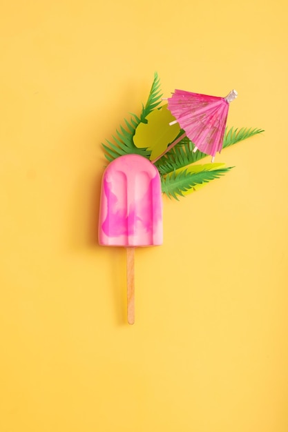 Pink ice cream with palm leaves and umbrella on a yellow background Minimalist creative summer composition top view