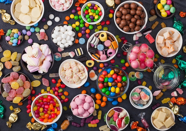 Pink lollipop candies in jar with various milk chocolate and jelly gums candies on black with liquorice allsorts and strawberry bonbons and large variety of sweets and candies