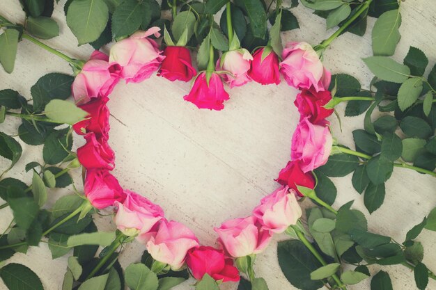 Pink and magenta fresh roses frame in heart shape on wooden aged background, retro toned