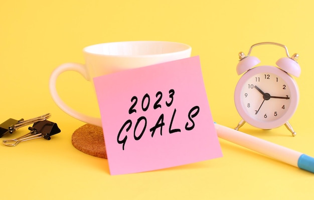 Pink paper with the text  goals on a white cup clock pen on a yellow background