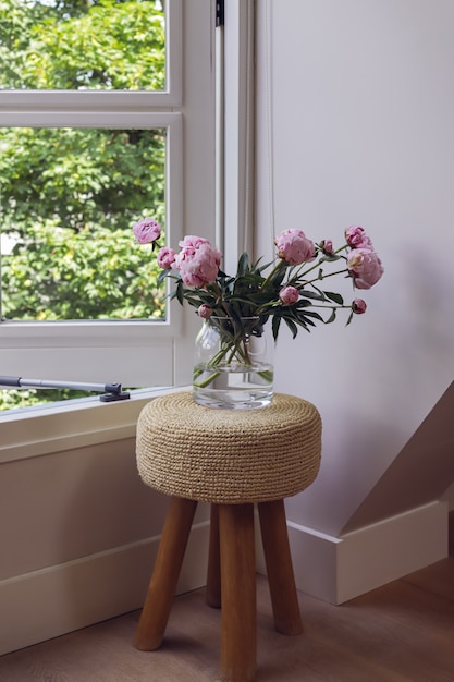 Pink peony bouquet in glass vase standing on small stool next to open window on green  background. 