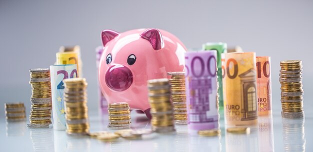 Pink piggy bank in the middle of rolled euro banknotes and towers with coins.