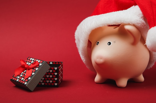 크리스마스 모자가 있는 분홍색 돼지 저금통, 빨간색 배경에 격리된 선물 리본이 있는 선물 상자. 돈 축적, 투자, 부 개념입니다. 새해 복 많이 받으세요, 생일 휴가. 복사 공간을 비웃습니다.
