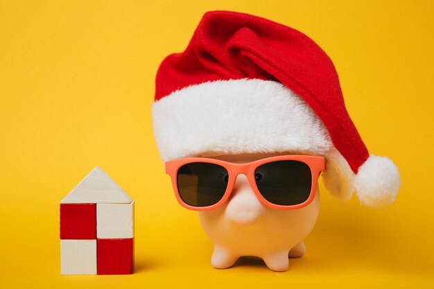 Pink piggy money bank with sunglasses christmas hat, toy wooden house isolated on yellow wall background. money accumulation investment banking services wealth concept. copy space advertising mock up