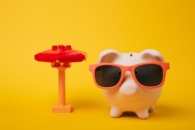 Pink piggy money bank with sunglasses, toy umbrella isolated on yellow wall background. Money accumulation investment, banking or business services, wealth concept. Copy space advertising mock up.