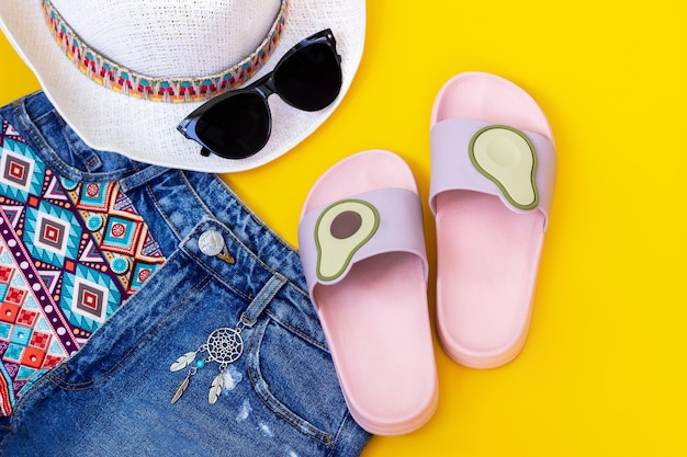 Pink and purple female beach flipflops with avocado halves denim shorts with a boho pattern and a dream catcher black sunglasses and white hat isolated on a color yellow wall