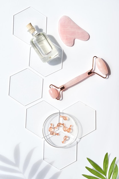 Pink quarts face roller, gua sha stone, serum capsules in chemical glass dish, hexagons, palm leaves. Beauty facial massage. Natural cosmetics laboratory. Off white top view in green and pink.