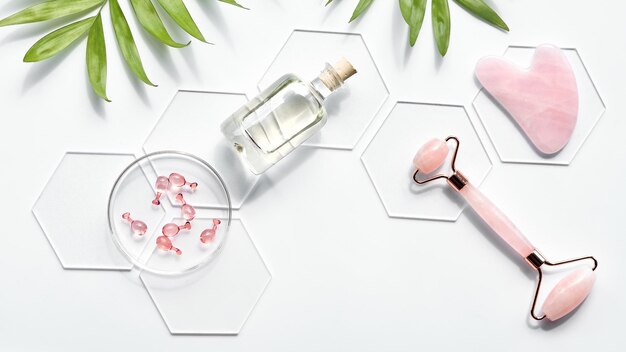 Pink Quarts stone face roller serum capsules in round glass dish hexagons palm leaves Gua sha stone for beauty facial massage therapy Natural cosmetics laboratory Off white flat lay text space