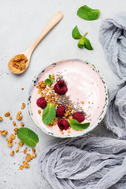 Ciotola per frullato di banana e lampone rosa con muesli, semi di chia, cocco e menta su cemento grigio chiaro.