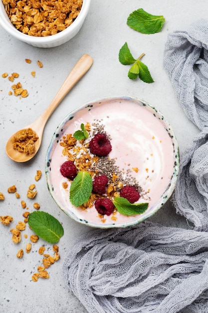 Pink raspberry banana smoothie bowl with granola, chia seeds, coconut and mint on light gray concrete.   