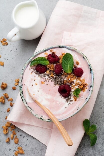 Pink raspberry banana smoothie bowl with granola, chia seeds, coconut and mint on light gray concrete.   