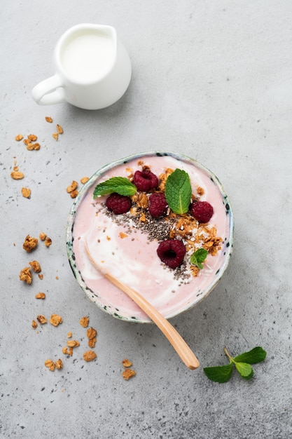 Ciotola per frullato di banana e lampone rosa con muesli, semi di chia, cocco e menta su cemento grigio chiaro.