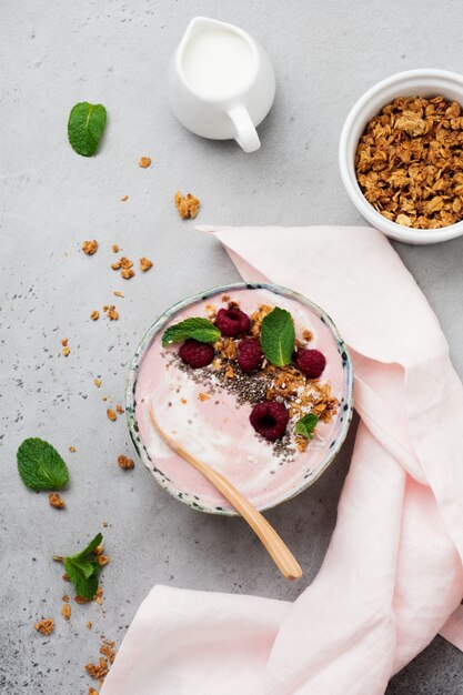 Pink raspberry banana smoothie bowl with granola, chia seeds, coconut and mint on light gray concrete.   