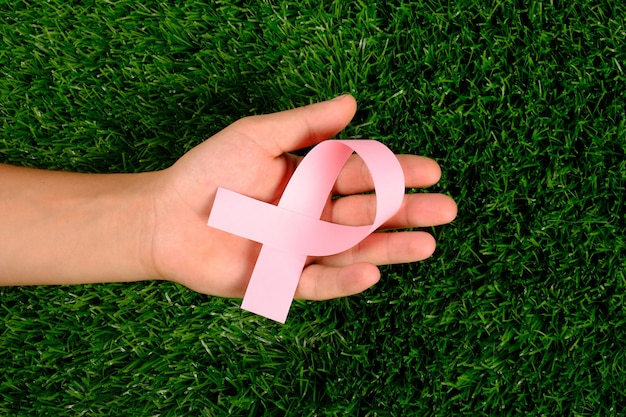 Pink ribbon in hand a symbol of the fight against cancer on green grass.