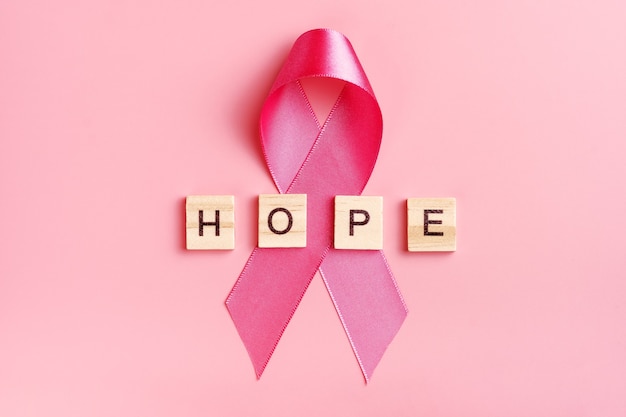 Pink ribbon and wooden letters with word hope on pink background.