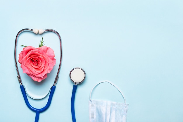 Pink rose heart, stethoscope and protective mask on blue background. Thank you doctor and nurse day concept.