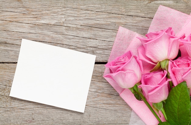 Букет розовых роз и пустая открытка за деревянным столом