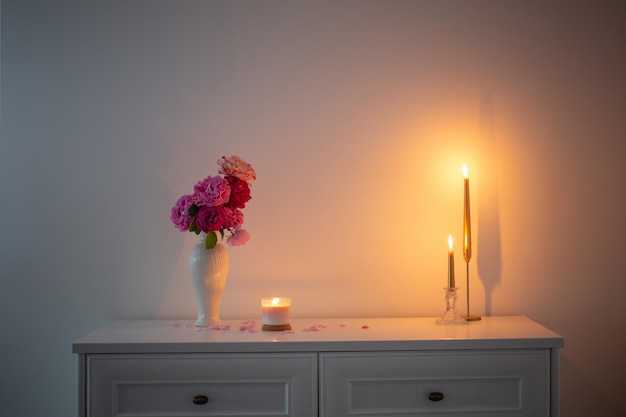 Pink roses in vase and burning candle on background white wall