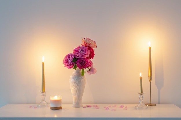 Pink roses in vase and burning candle on background white wall