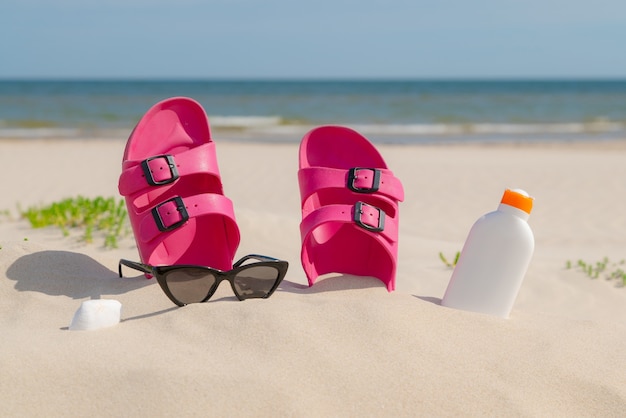 Photo pink sandals, sunglasses and sunscreen at the beach on a beautiful sunny day.