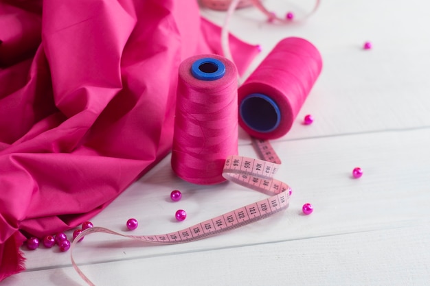 Pink satin fabric with thread and centimeter on wooden background with beads.